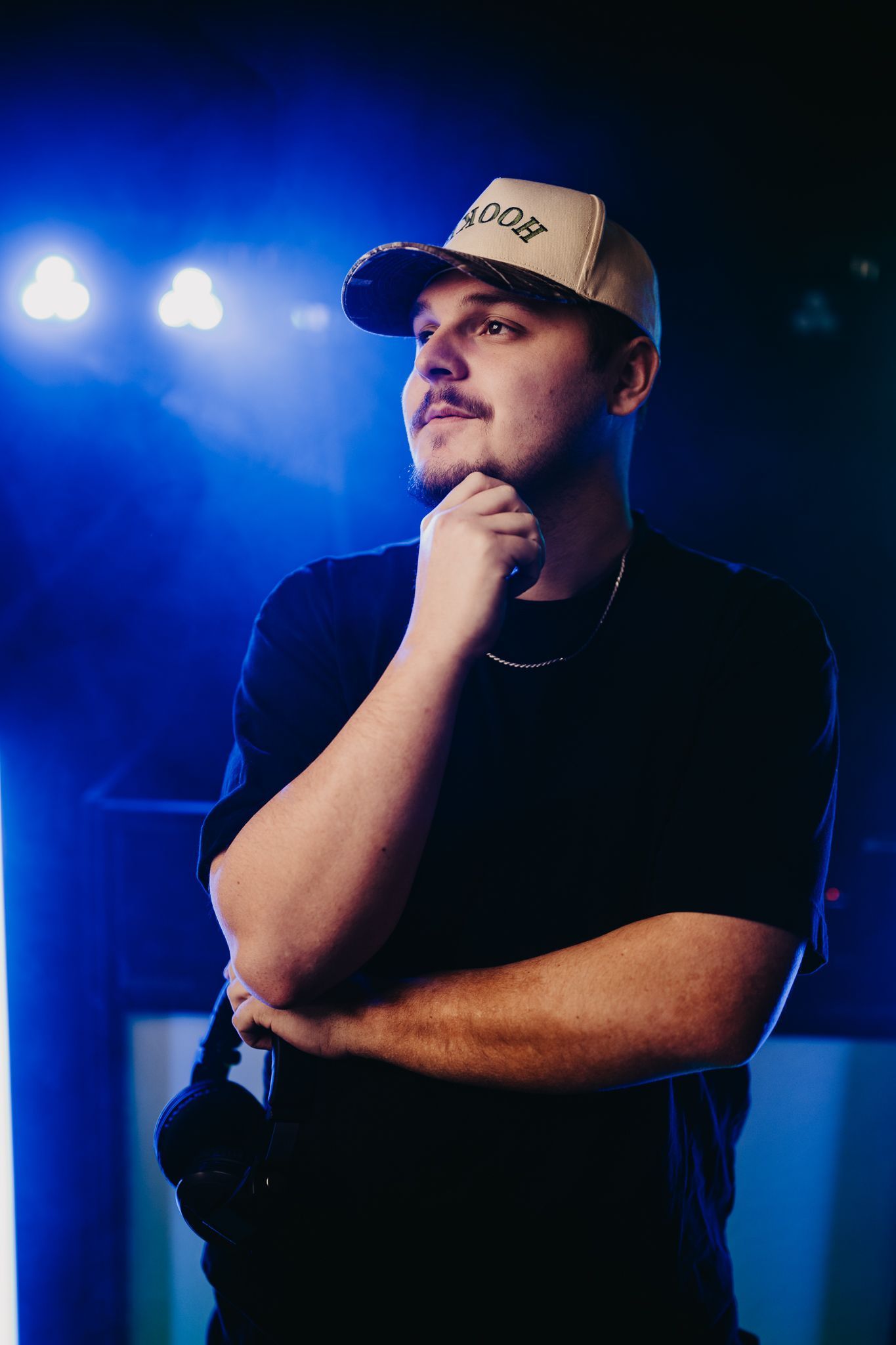A person in a baseball cap stands under blue stage lighting, looking to the side with their hand on their chin.
