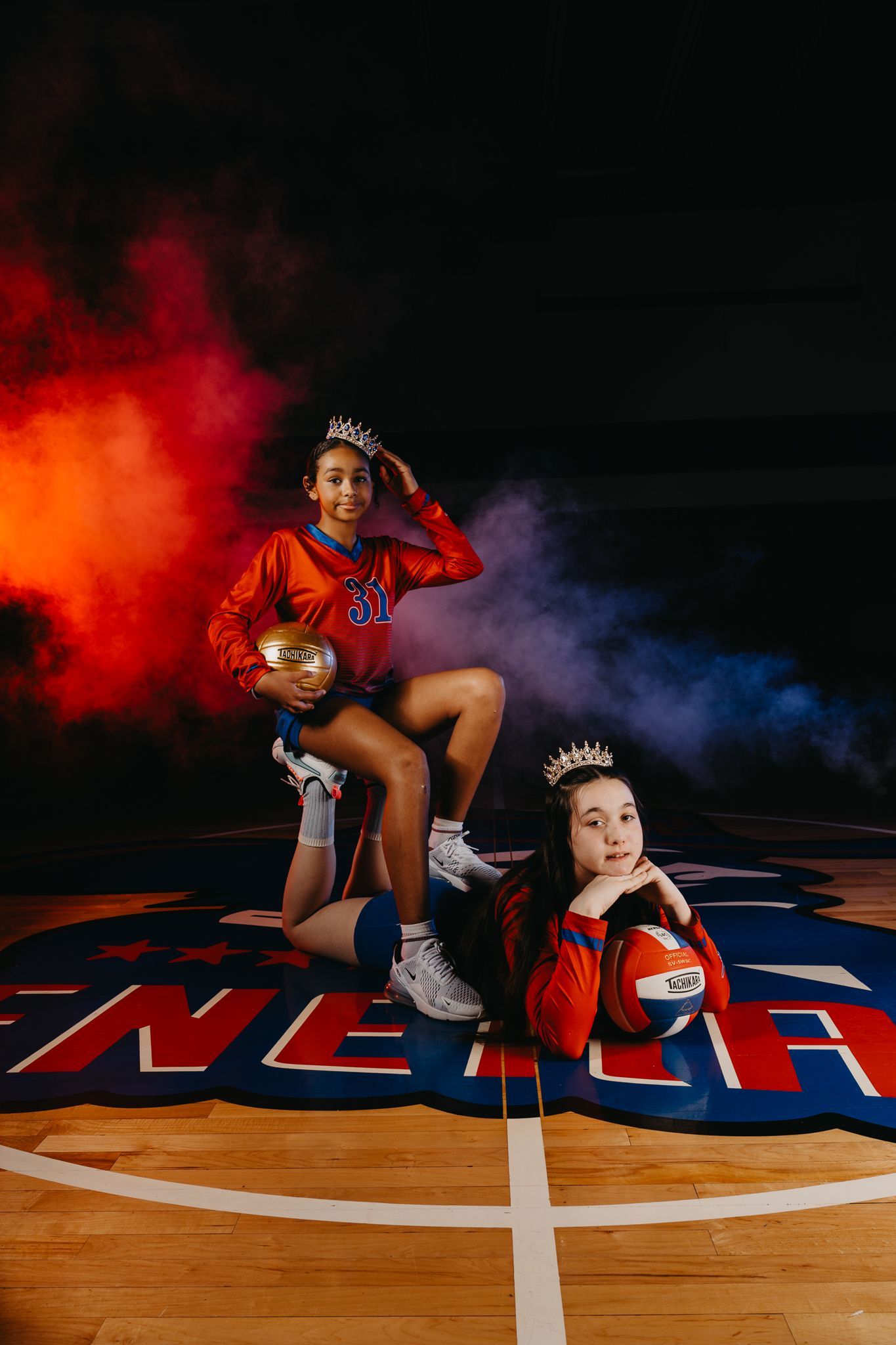 Two people wearing sports jerseys and crowns pose with basketballs on a gymnasium floor amid orange and blue smoke.