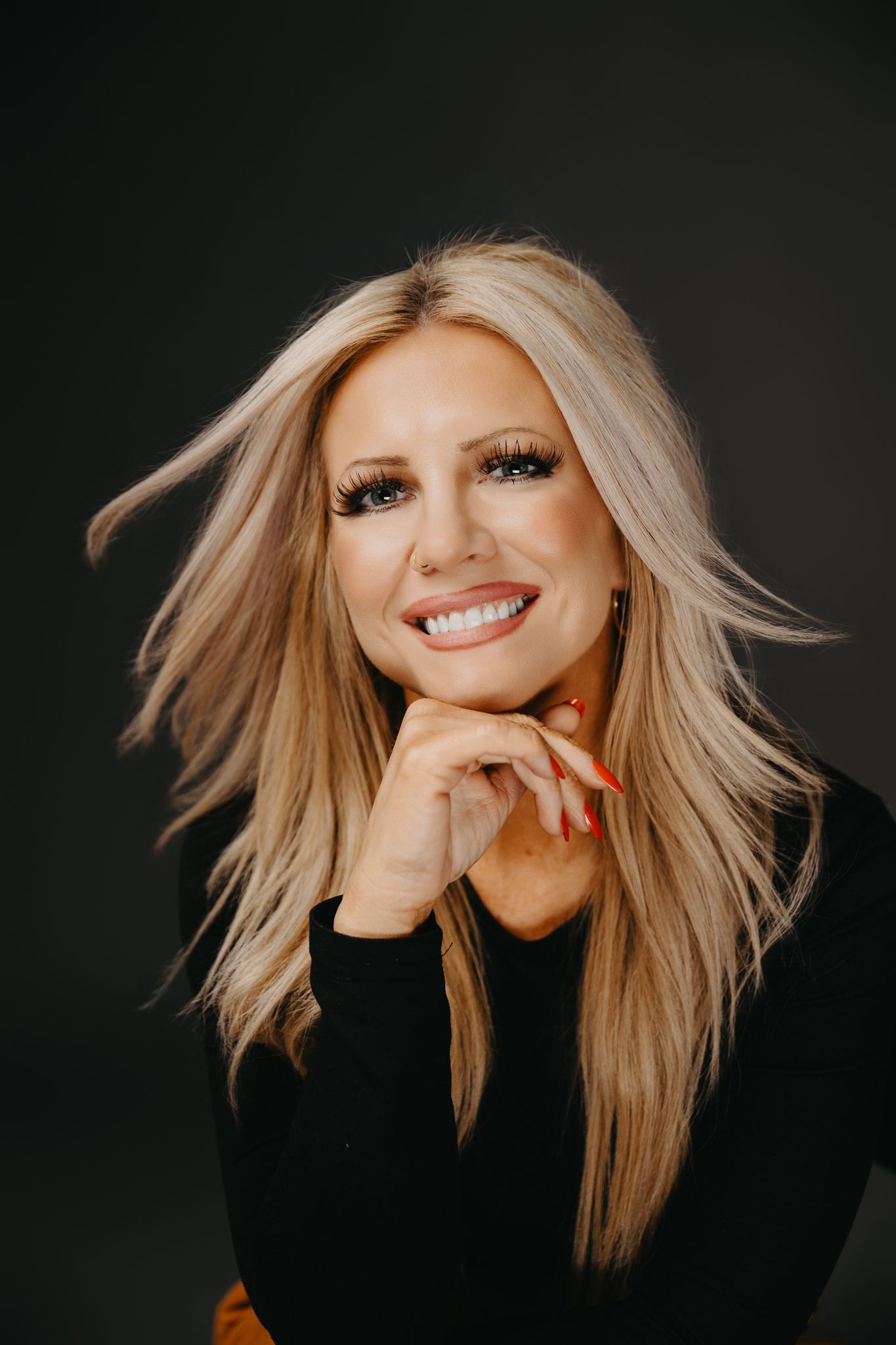 A blonde person with long, wavy hair smiles with their chin resting on their hand against a dark, neutral background.