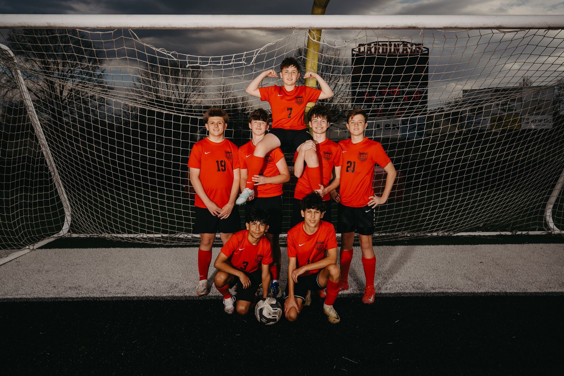 A group of eight soccer players in orange uniforms pose together in front of a soccer net on an outdoor field.