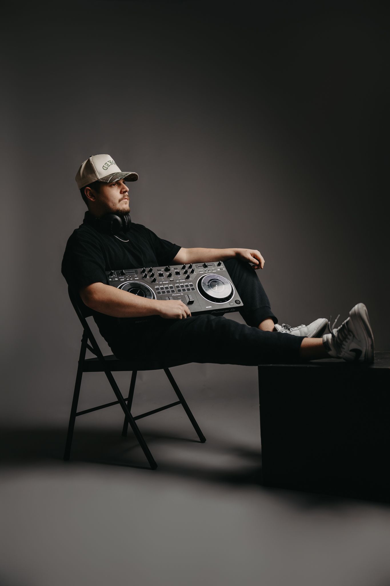 A person wearing a cap sits on a chair in a dark studio, holding a DJ controller on their lap with feet resting on a box.