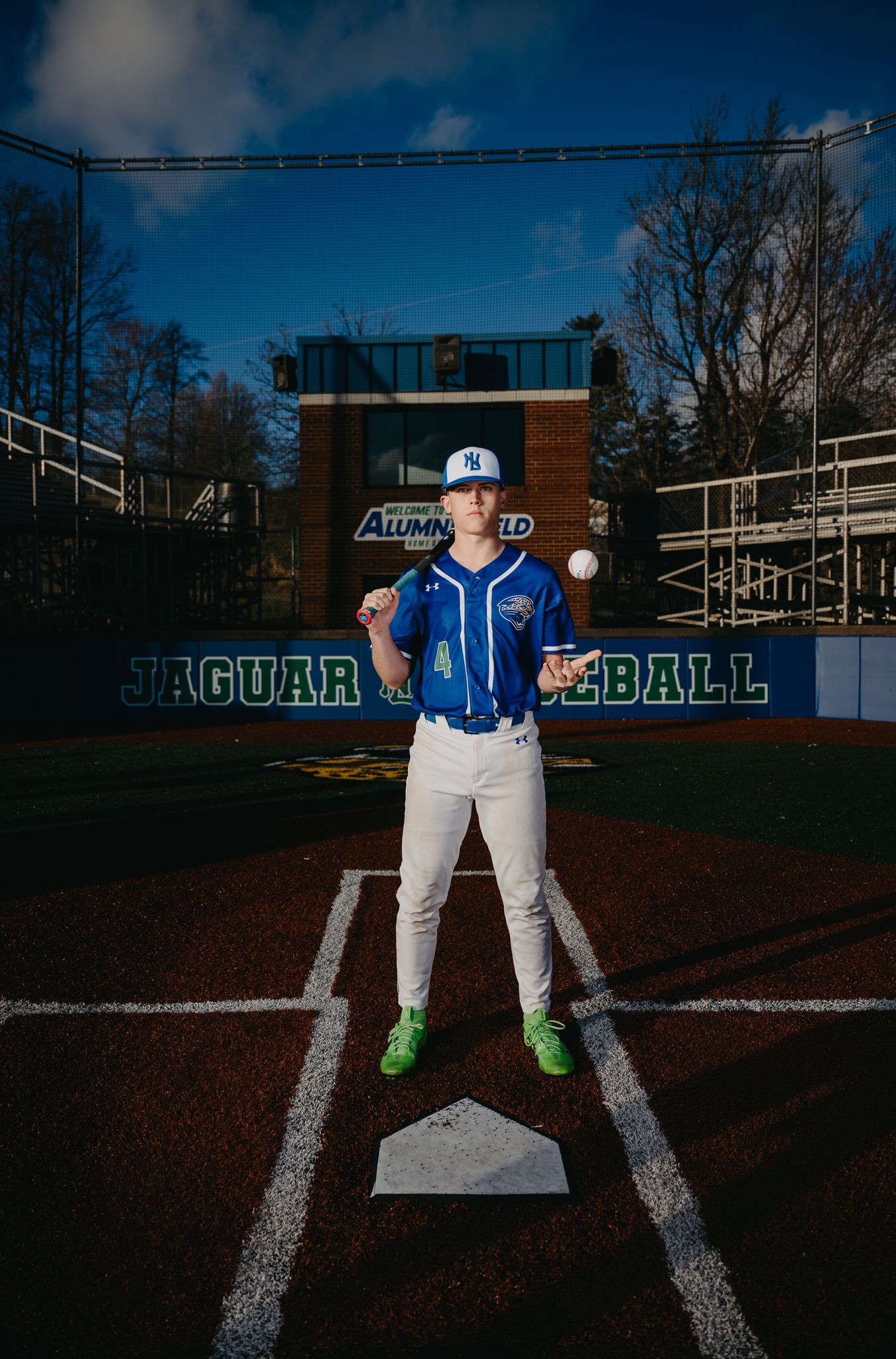 A baseball player in a blue uniform stands at home plate on a dirt field, holding a ball in each hand.