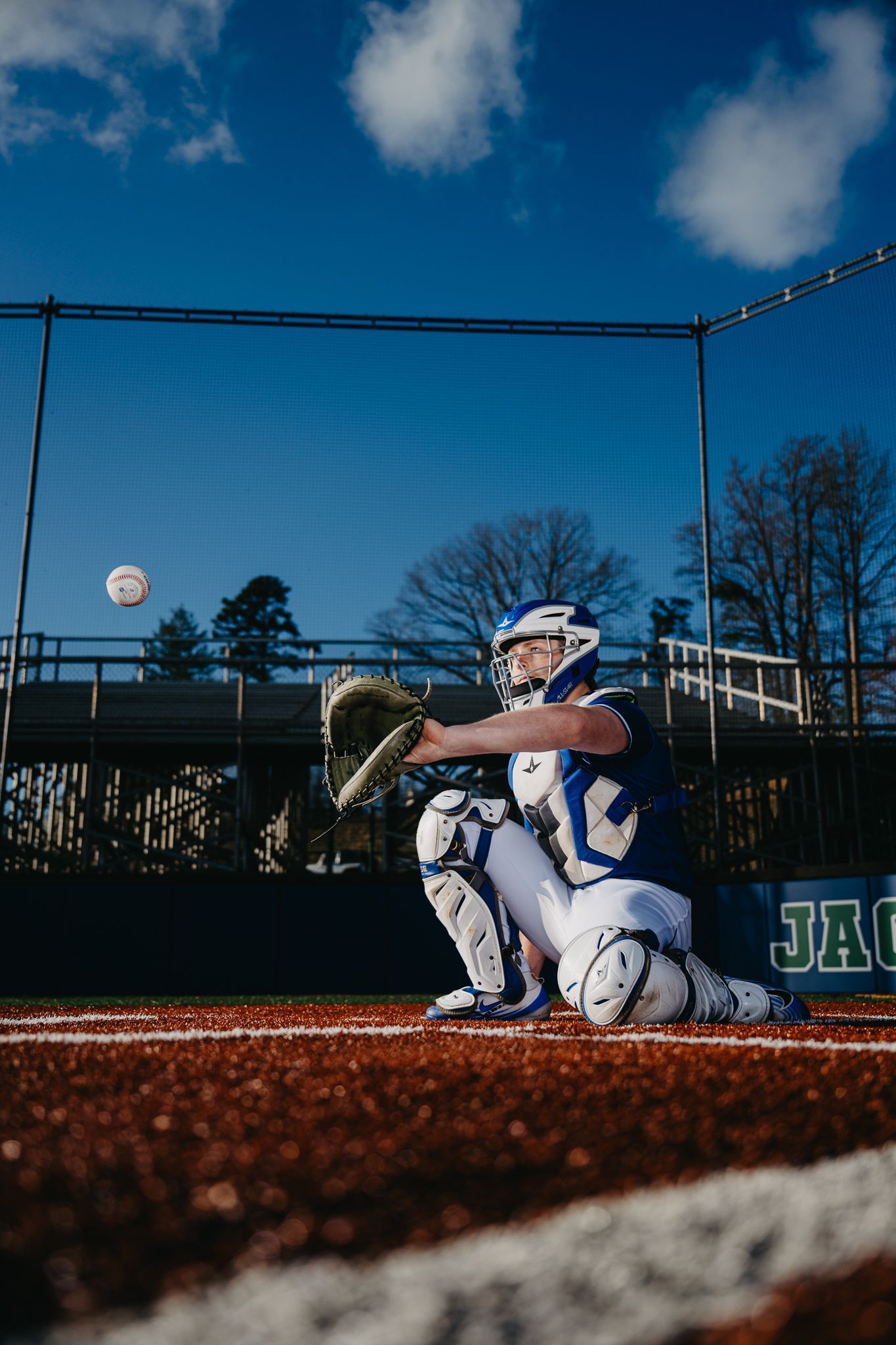 A baseball catcher in a blue and white uniform kneels on the field, reaching out to catch a ball mid-air.