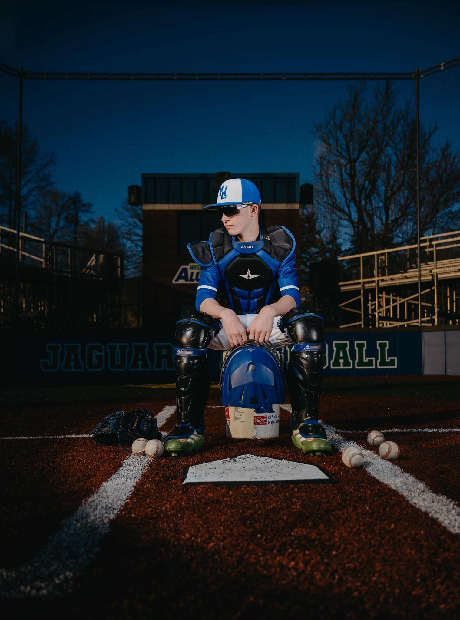 A baseball catcher in blue gear sits on a bucket at home plate on a baseball field at dusk.