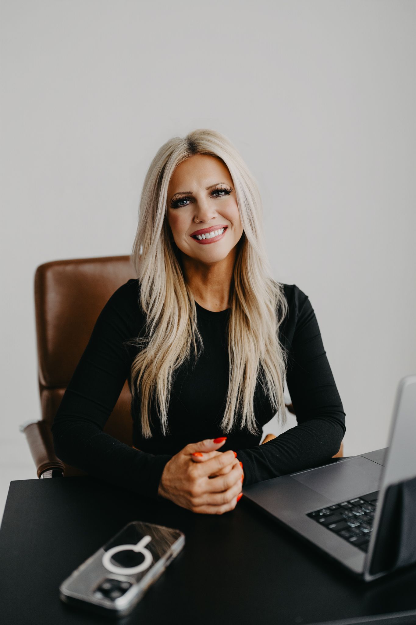 A person with long blonde hair sits at a desk, smiling with their hands clasped near a laptop and a smartphone.