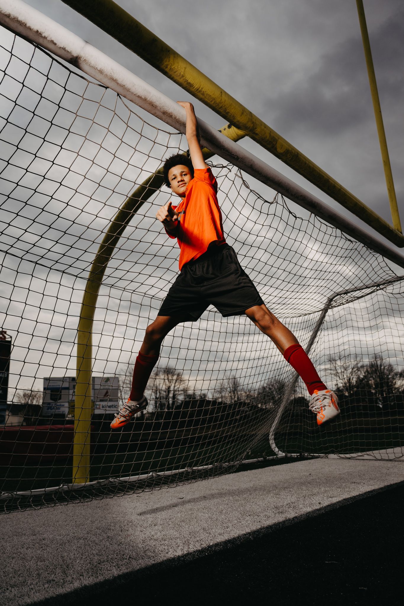 A person in an orange shirt and black shorts hanging from the crossbar of a soccer goal under a cloudy sky.