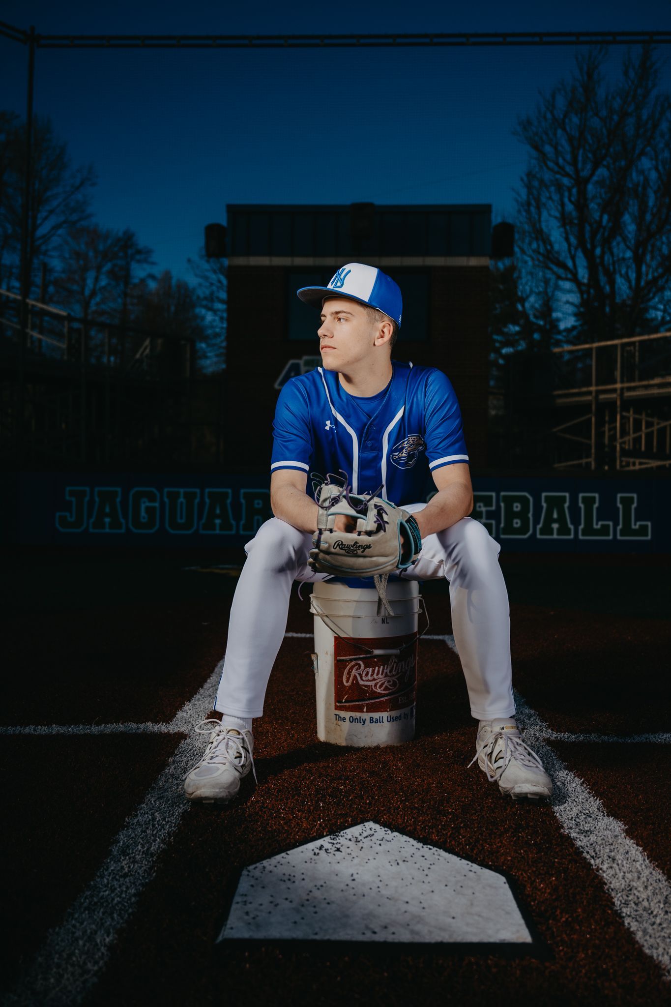 A baseball player in a blue uniform sits on a bucket behind home plate at a baseball field at dusk.