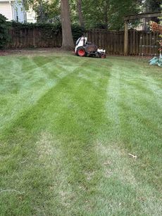 Lawn with freshly cut stripes. A small tractor with attachments sits in the background near a tree and fence.