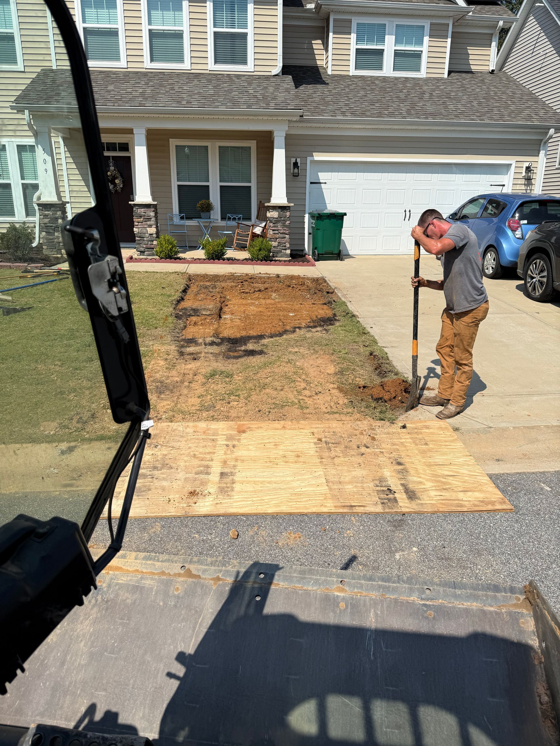 Man digging in front yard. Brown dirt, wooden pallets, gravel driveway, house in background.