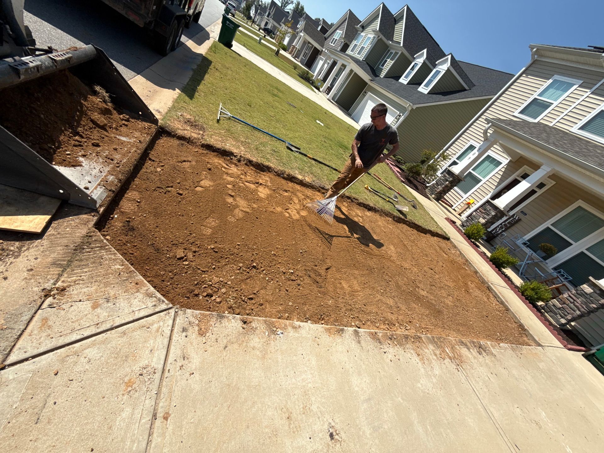Man standing in a dirt plot preparing for planting, surrounded by a concrete sidewalk and townhouses.
