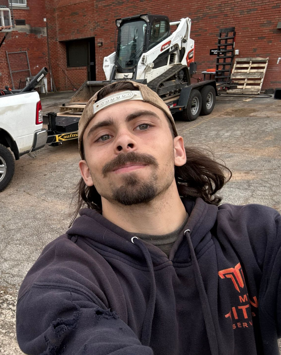 Man in cap and hoodie smiles, with a skid steer and trailer in the background.