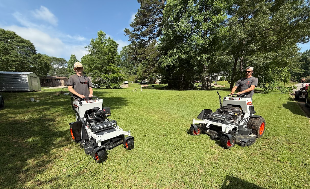 Two people operate small, grey and orange robotic mowers on a grassy lawn under a blue sky.
