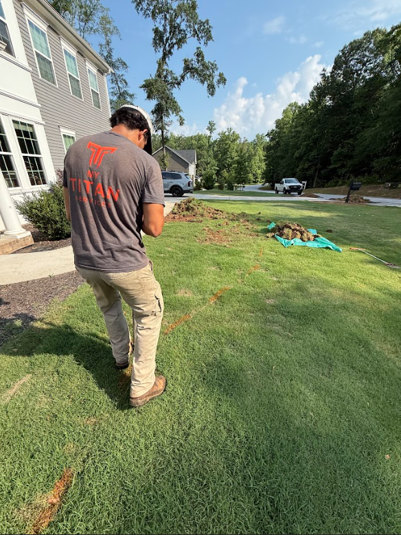 Man in work attire on grass, looking at device. House in background, small dirt area. Sunny day.