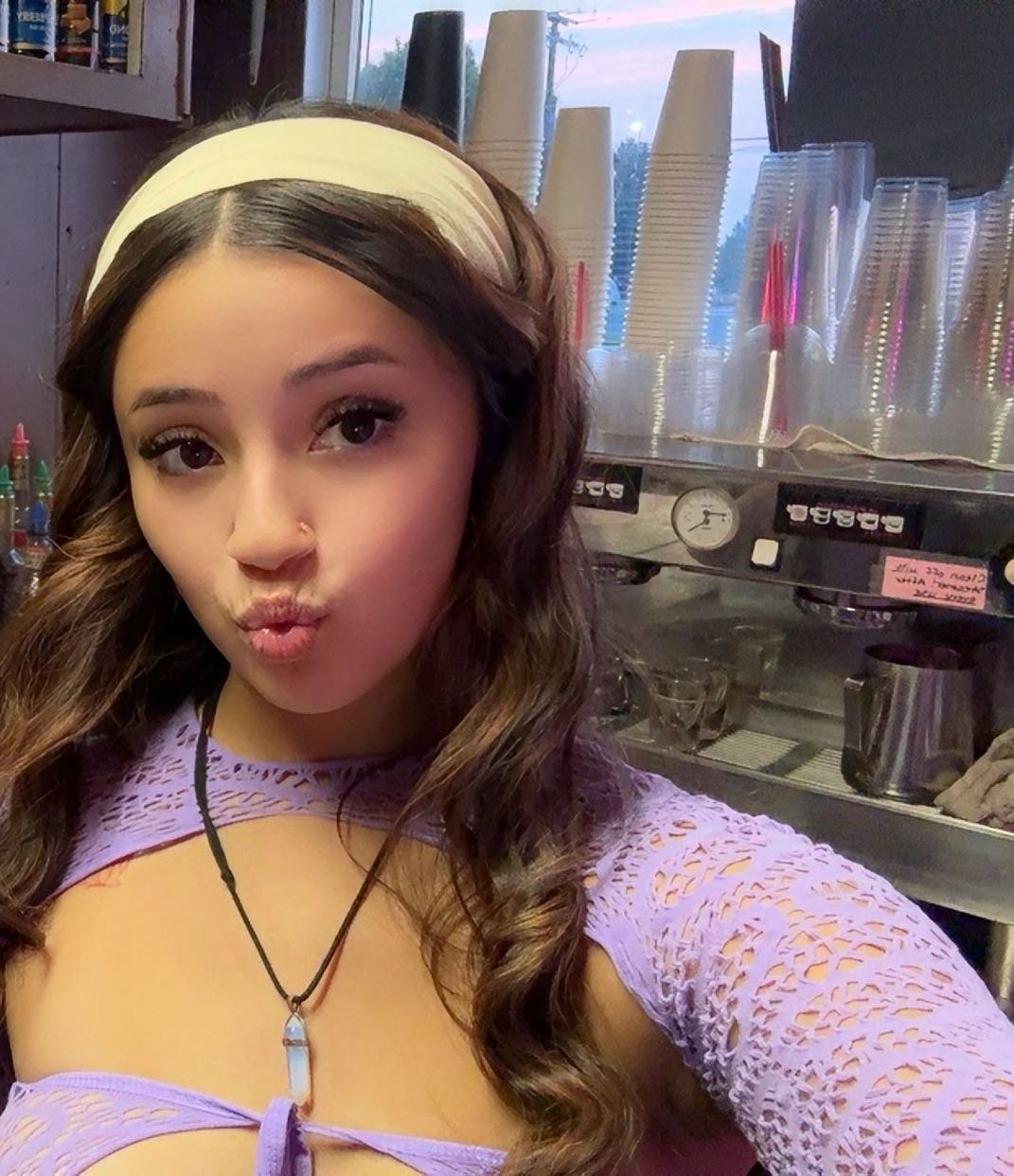 Woman with pout, wearing lavender top and headband, in coffee shop setting with espresso machine.