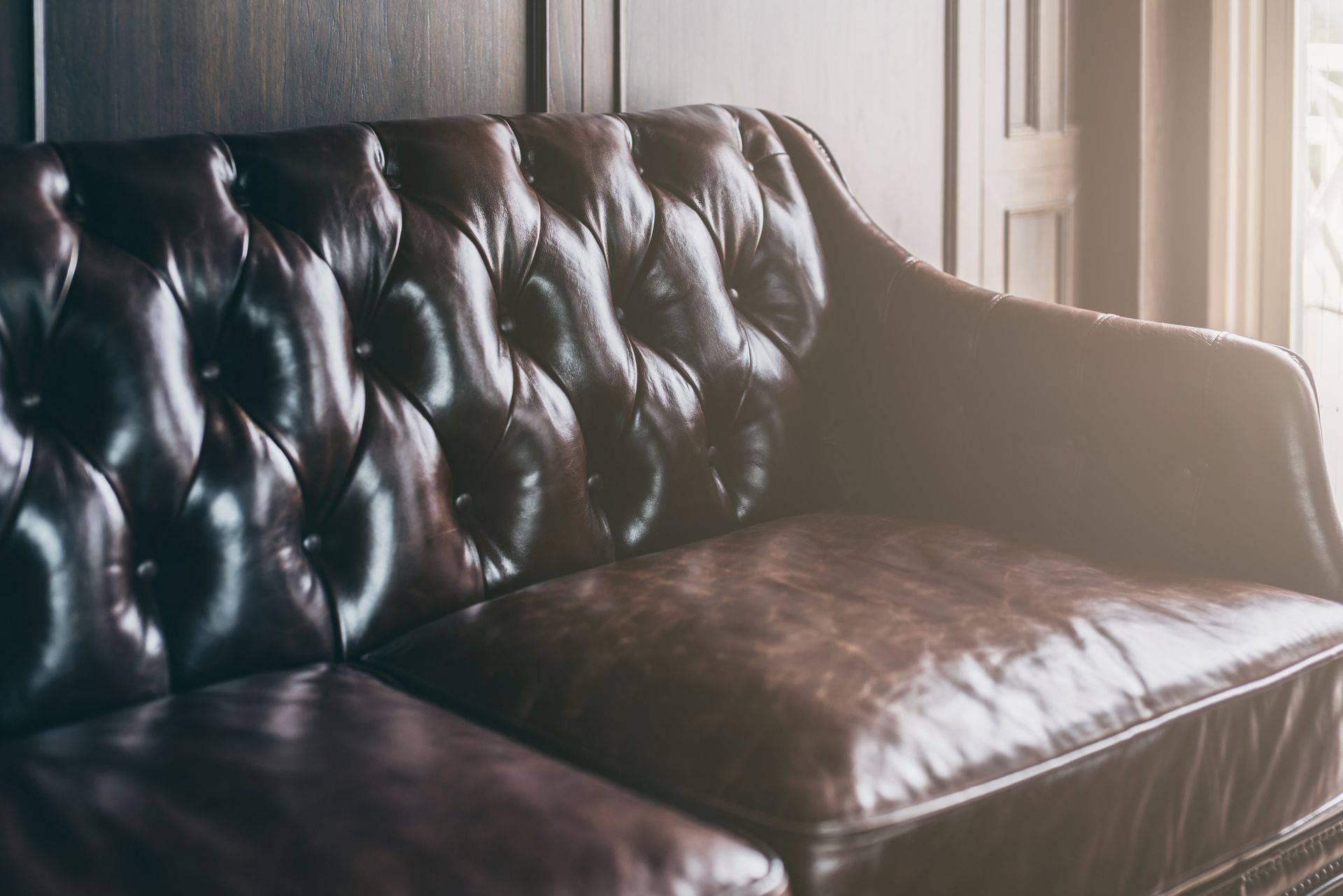Brown leather tufted sofa in front of dark wooden wall, lit by natural light.