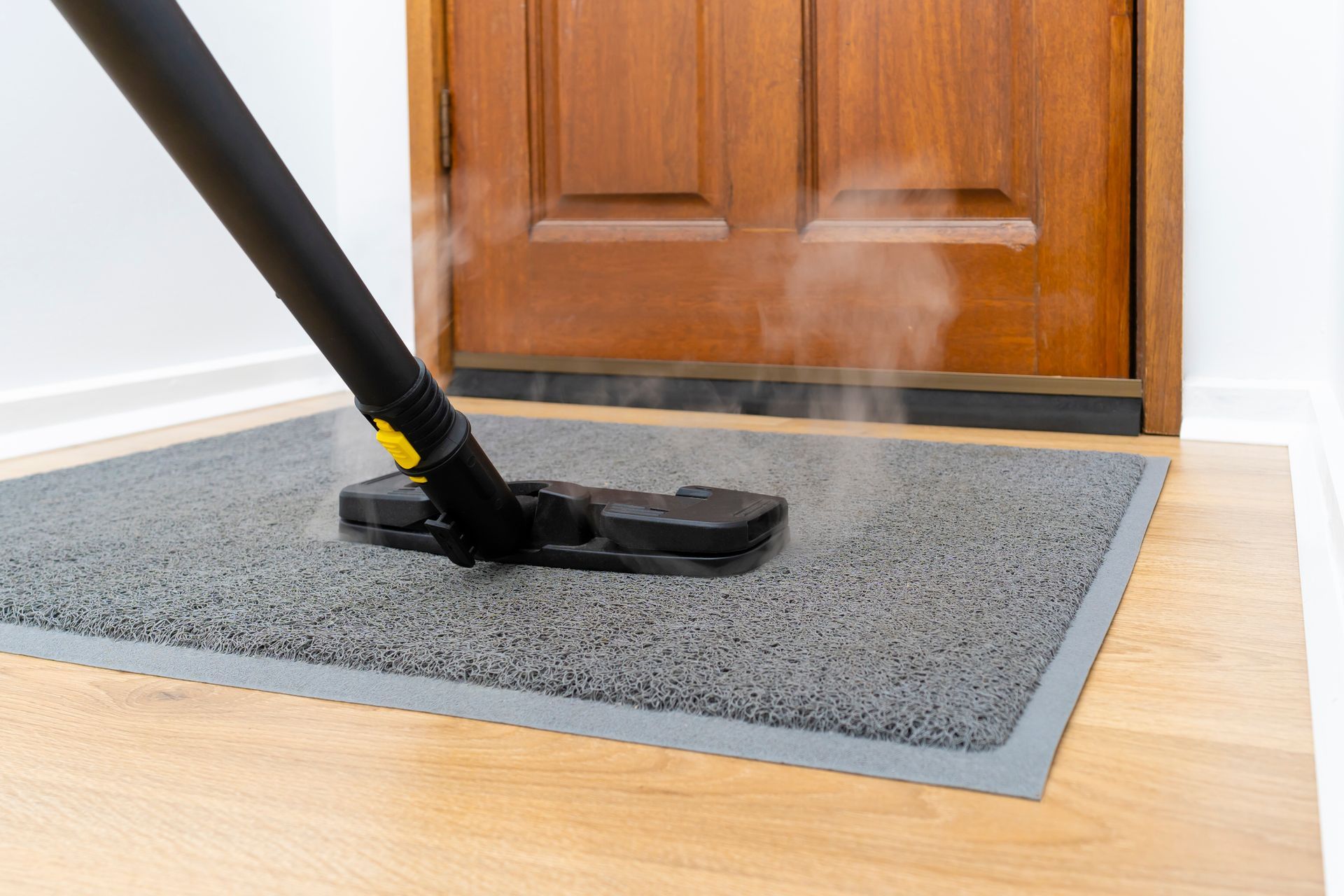 Steam cleaner sanitizing a gray door mat in front of a wooden door.