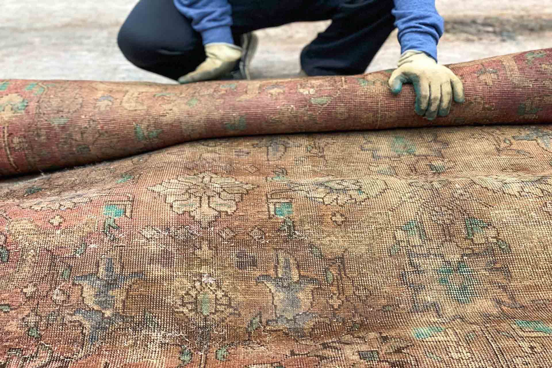 Person rolling up a faded, ornate rug on a light-colored surface. They wear gloves and a blue sweater.