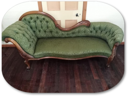 Green chaise lounge with button tufting and ornate wooden frame, on dark wood floor.