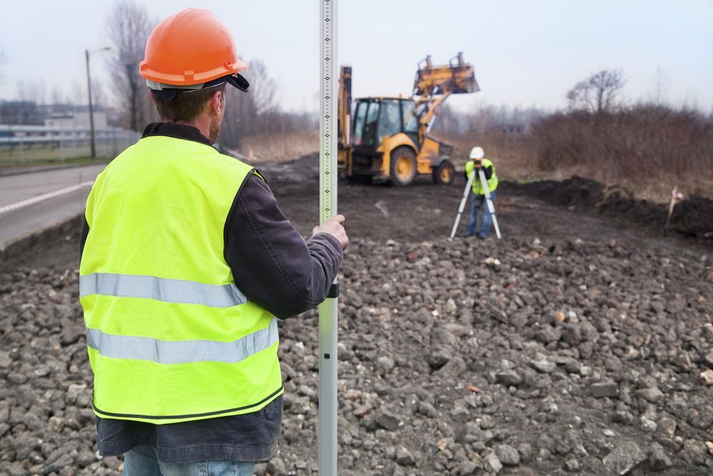 Man In A Yellow Vest Holding A Measuring Stick and Watching Bulldozer Digging — Surveyor In Young, NSW