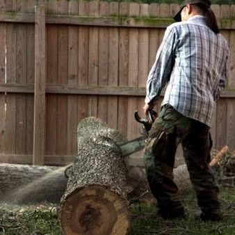 A person wearing a plaid shirt and camo pants uses a chainsaw to cut a log in front of a wooden fence.