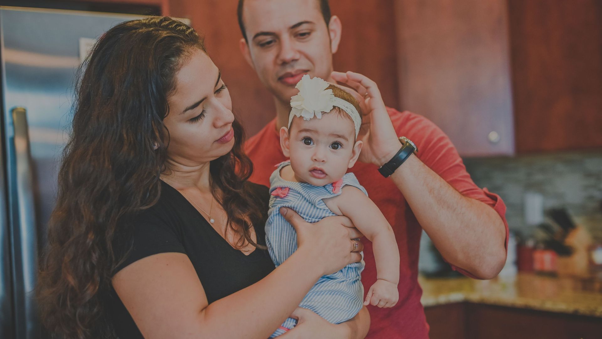 Unos padres ajustan la diadema de un bebé en una cocina. El bebé mira directamente al espectador.