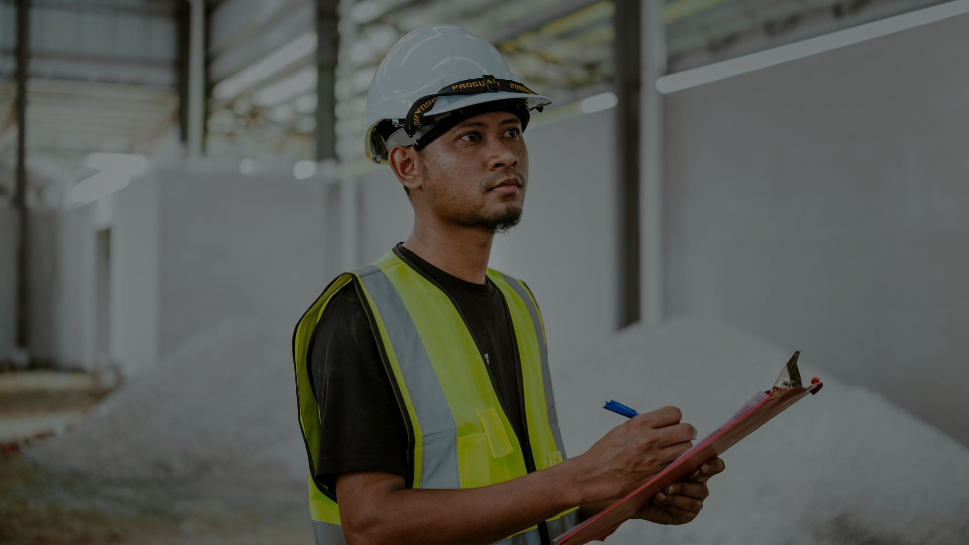 Un trabajador que lleva casco y chaleco de seguridad inspecciona un almacén, mientras escribe en un portapapeles.