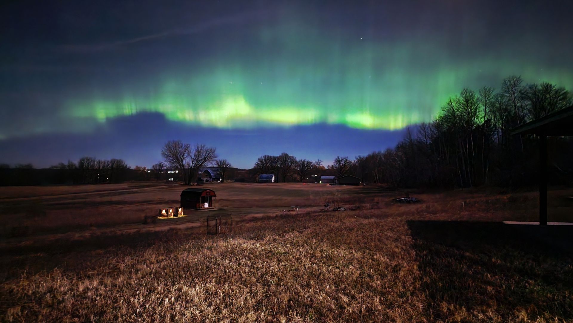 A vibrant green aurora borealis illuminates the night sky above a dark, quiet field with scattered trees and distant lights.