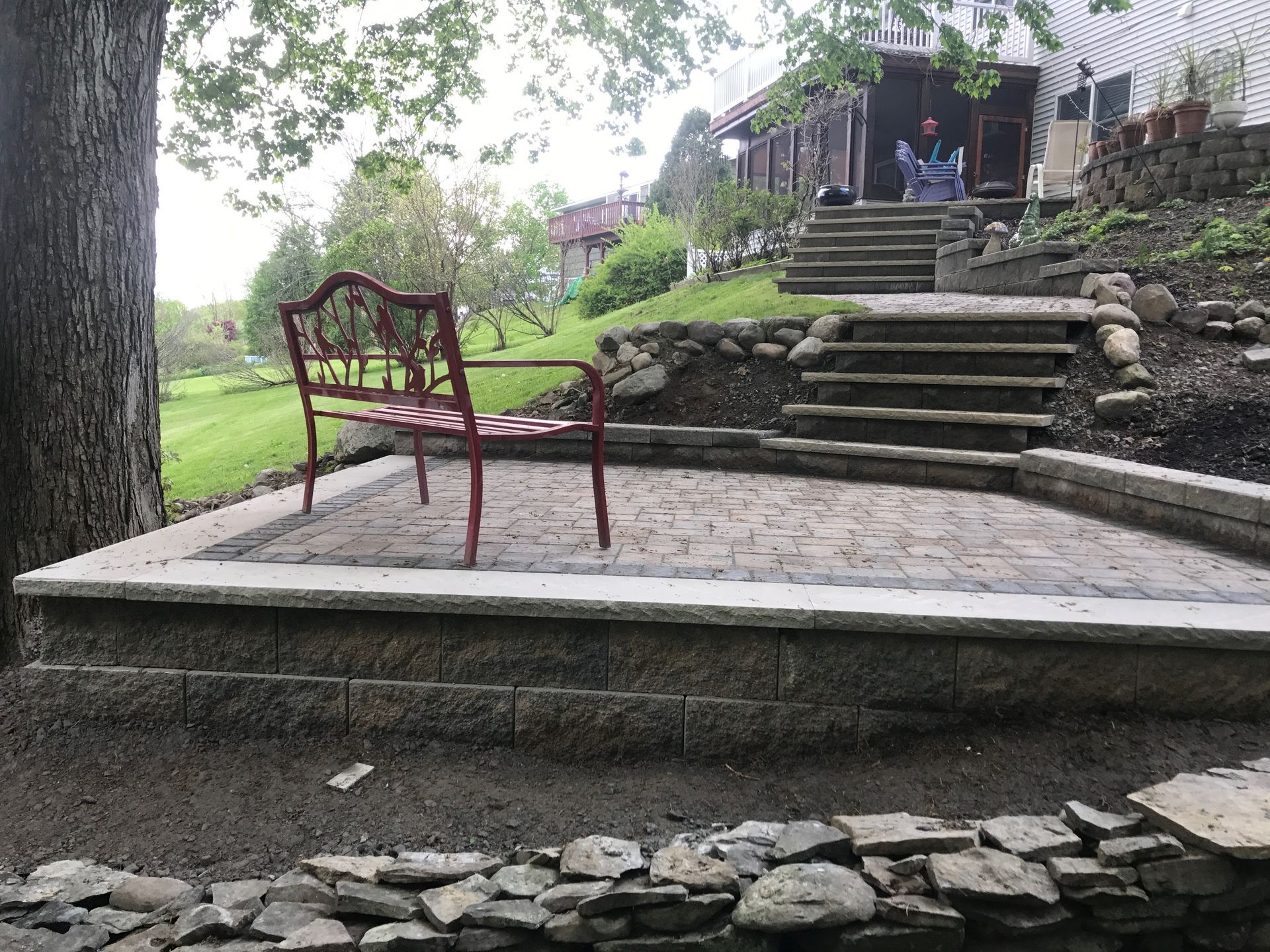 A red bench is sitting on a stone patio next to a tree.