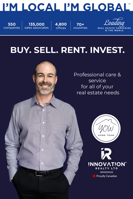 Man with a shaved head and beard, smiling, wearing a patterned shirt and black pants, hands in pockets.