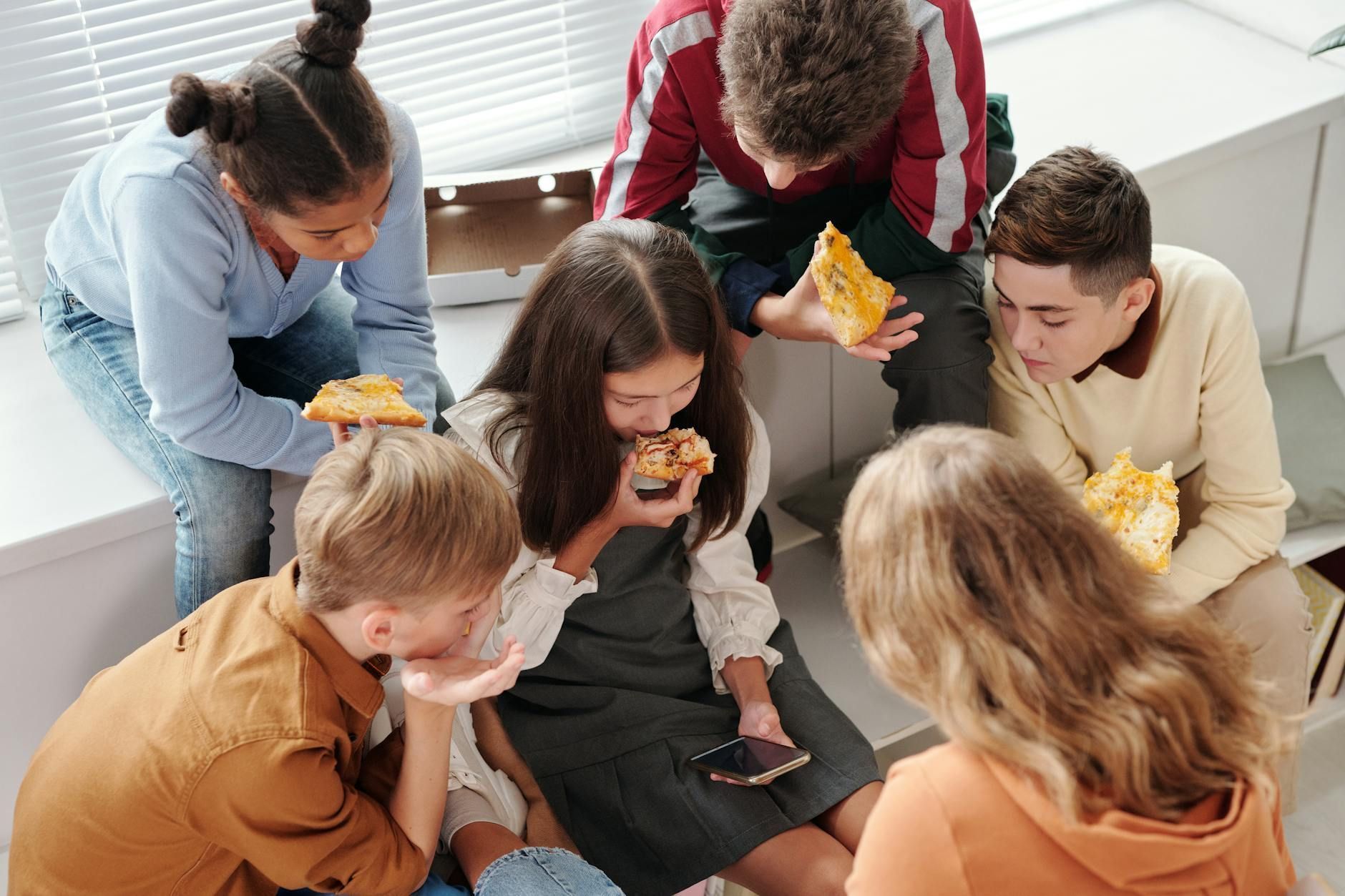 A group of young people are sitting near a window sill eating pizza.