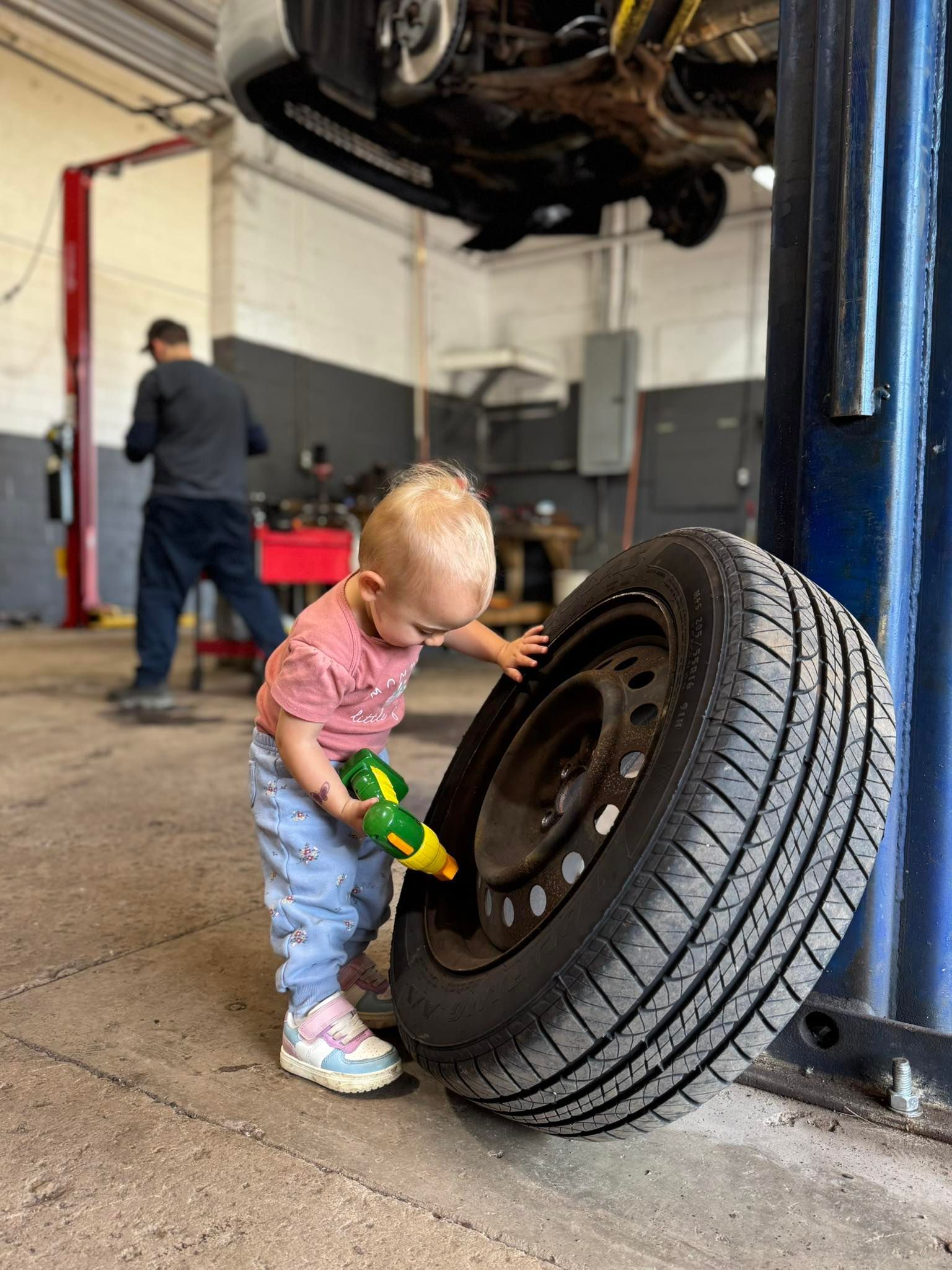 Toddler in a pink shirt inspecting a large tire in an auto repair shop | Medina’s Ace Auto