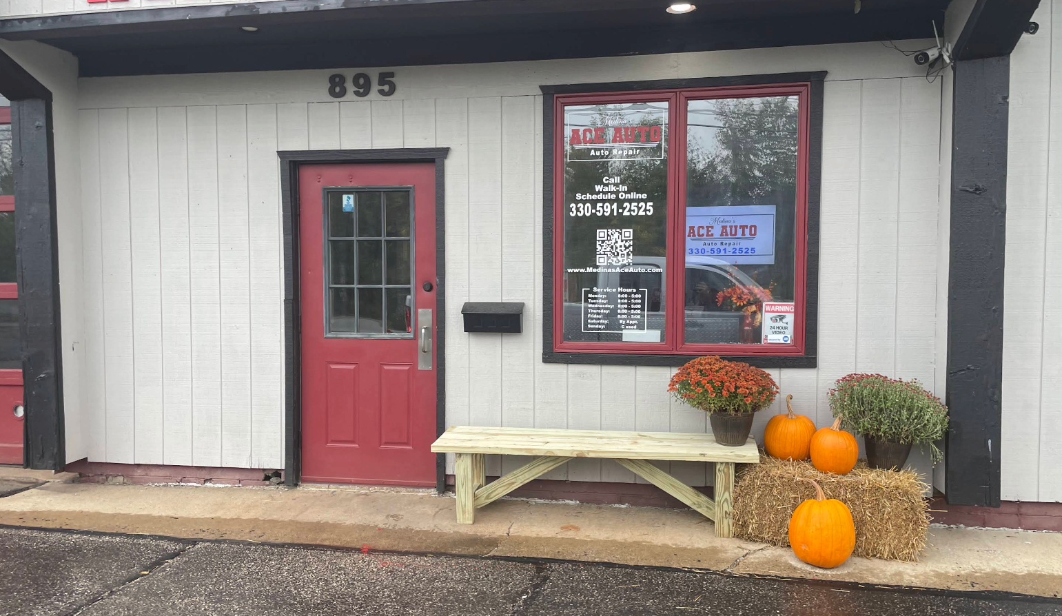 Small storefront with red door, bench, pumpkins, and window signage on a wet sidewalk. | Medina’s Ace Auto