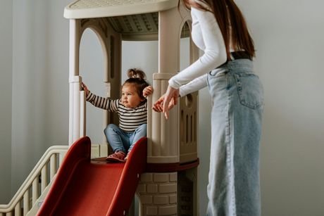 A woman is helping a little girl climb a slide in a playhouse.