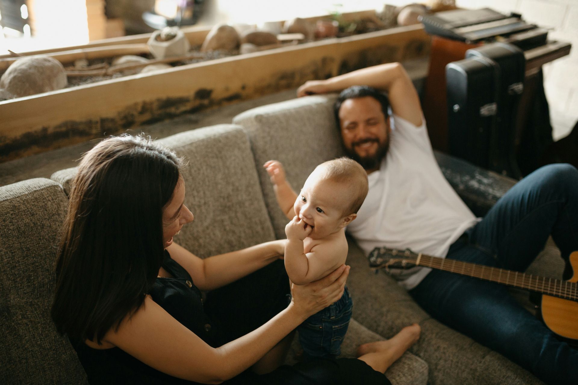 A man is playing a guitar while a woman holds a baby on a couch.