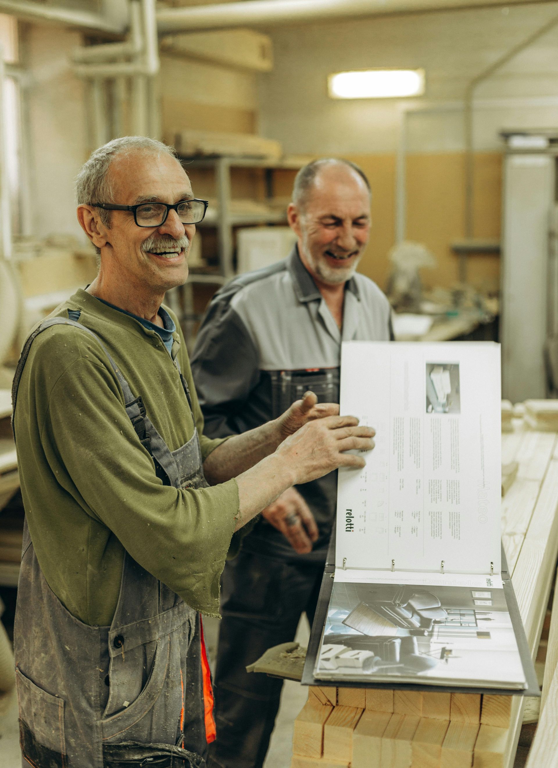 Two men are standing next to each other in a workshop holding a piece of paper.