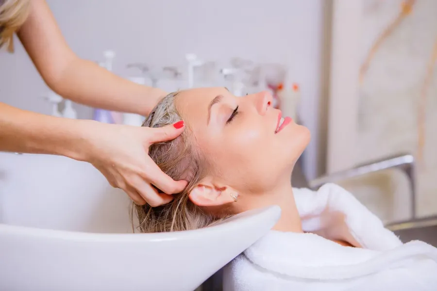 Woman reclining in a salon sink, hair being washed by a person's hands. She has her eyes closed and a relaxed expression.
