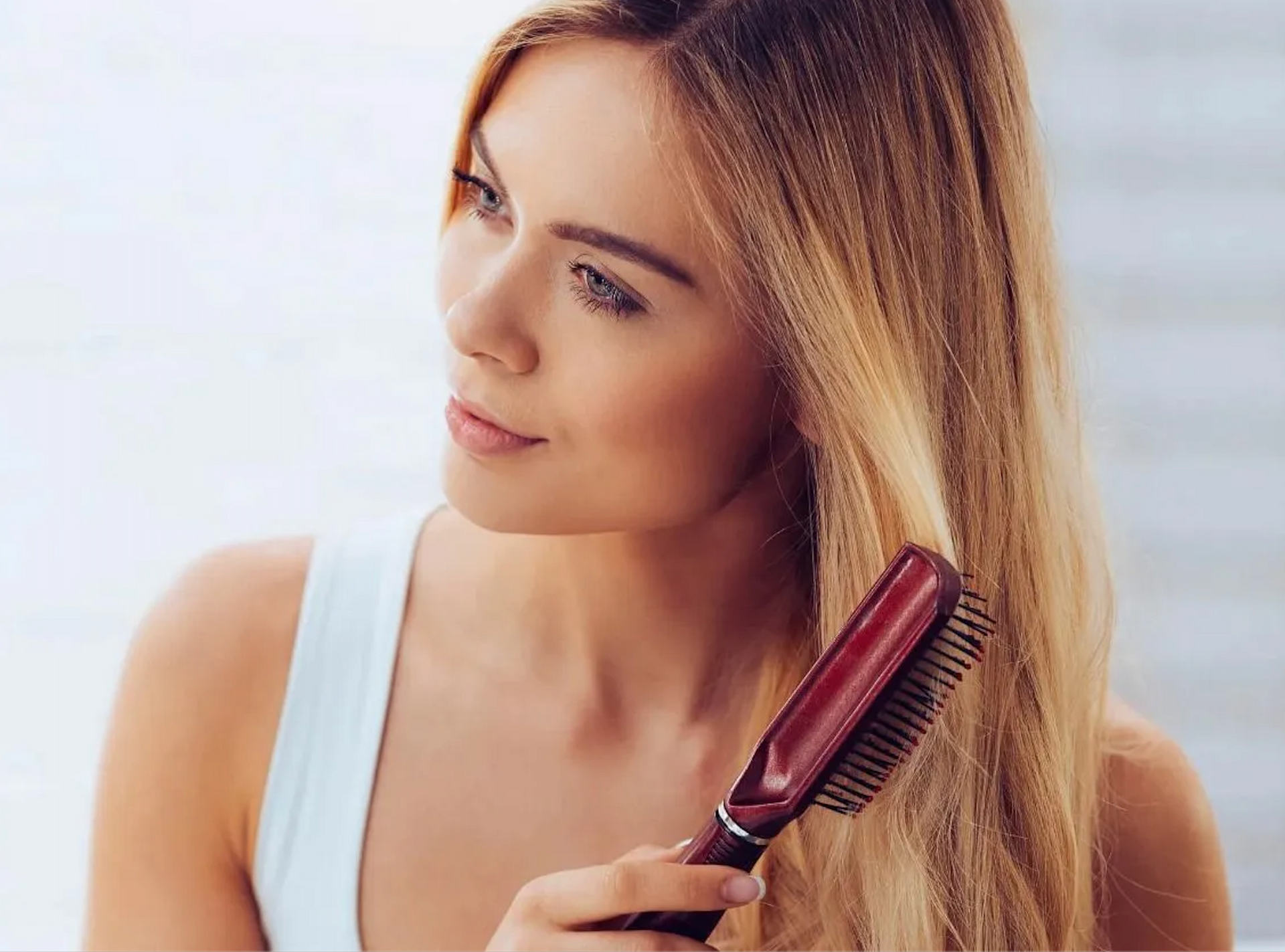 Woman with blonde hair, wearing a white tank top, brushes her hair with a red brush in a bright setting.
