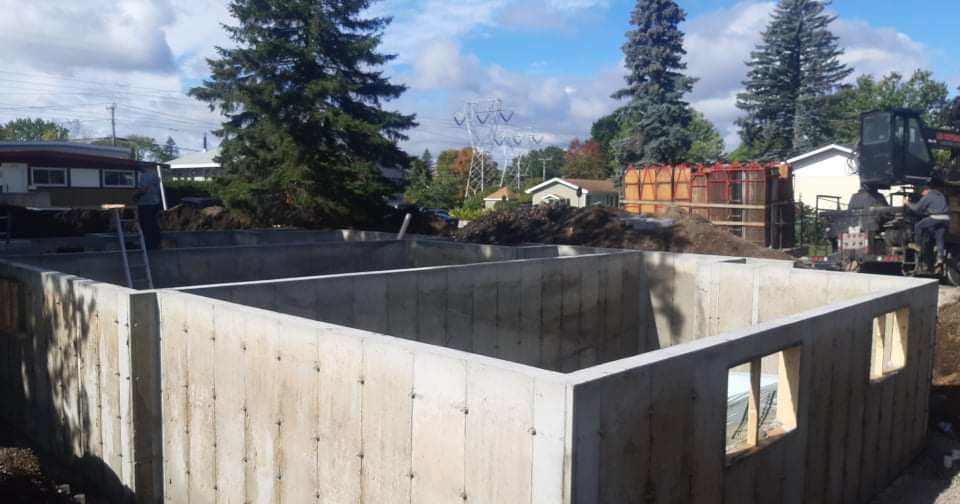 Fondations en béton d'un bâtiment en construction, fenêtres ouvertes, par une journée ensoleillée. Une pelleteuse et d'autres bâtiments sont visibles en arrière-plan.