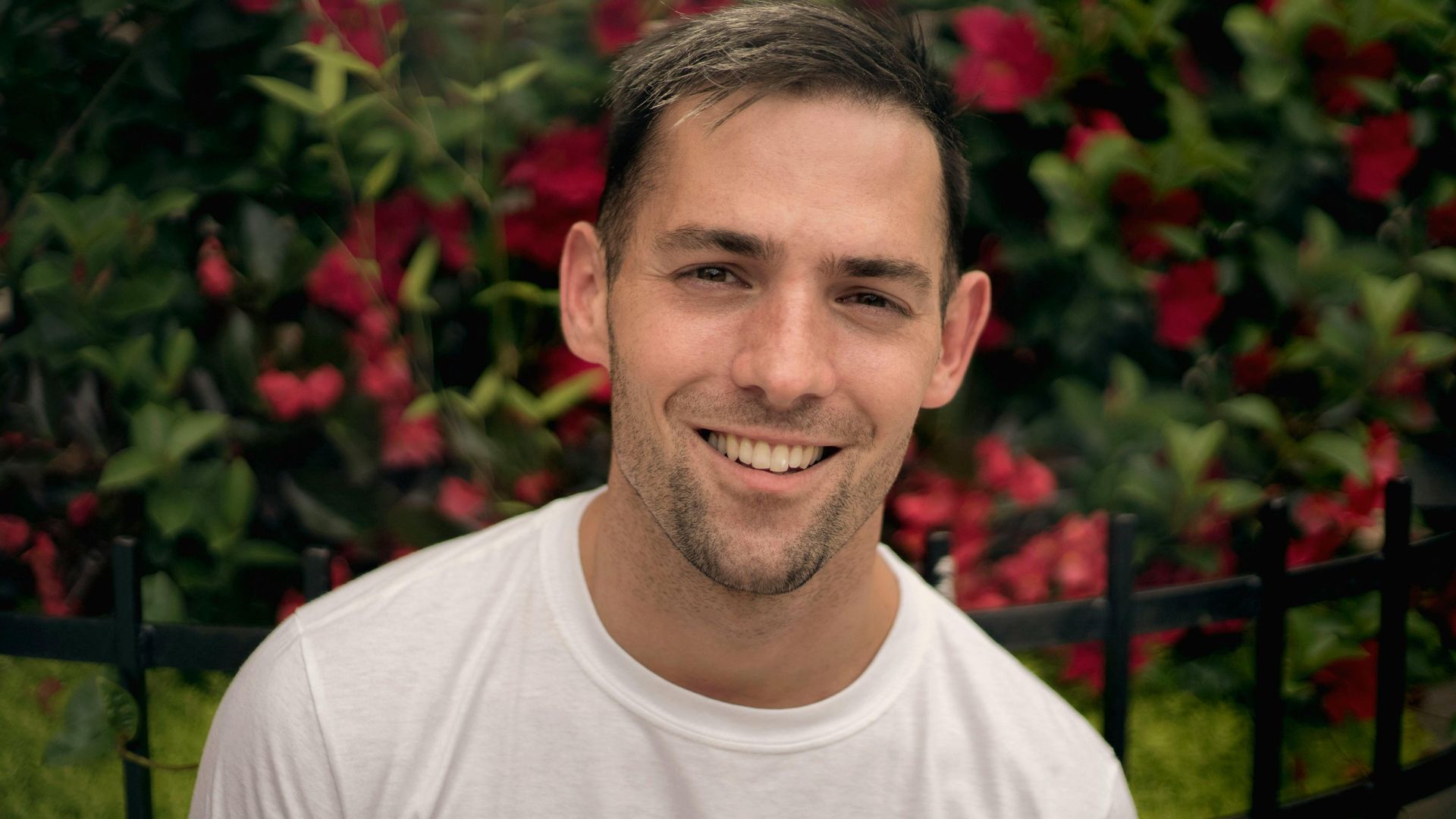 A man in a white shirt is smiling in front of a fence and flowers.
