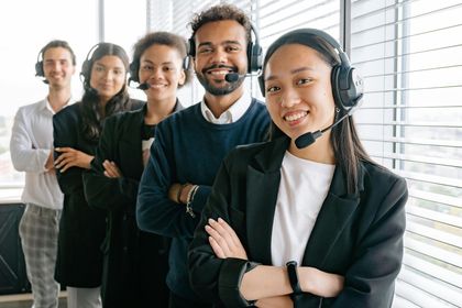 A diverse team of customer service representatives wearing headsets, smiling, and standing with arms crossed in an office setting.