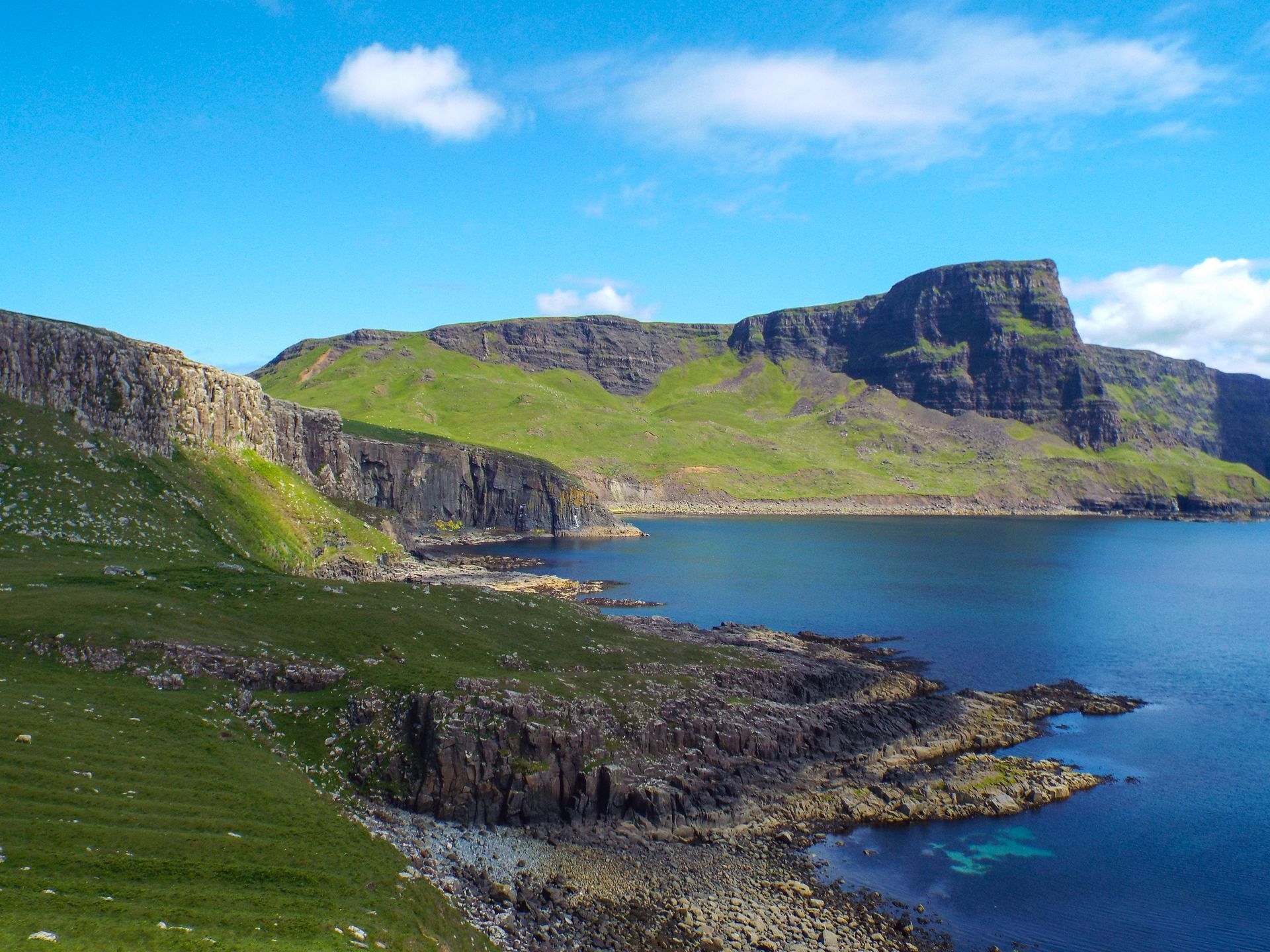 Coastal view of rocky cliffs, green hills, and dark blue water under a bright blue sky.