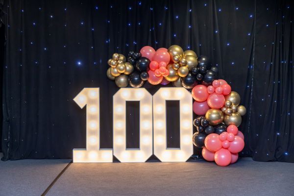 The number 100 is surrounded by balloons and lights.