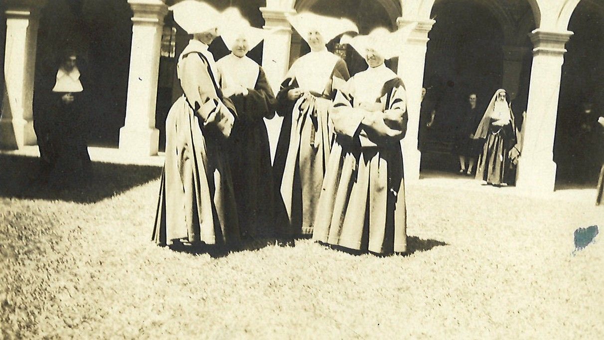 Group of people in long robes standing under stone arches in a courtyard, with one person in the background.