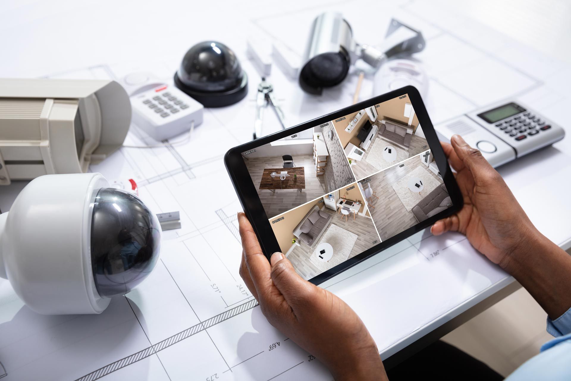 Hands holding a tablet showing multi-camera home security footage beside CCTV cameras and a keypad
