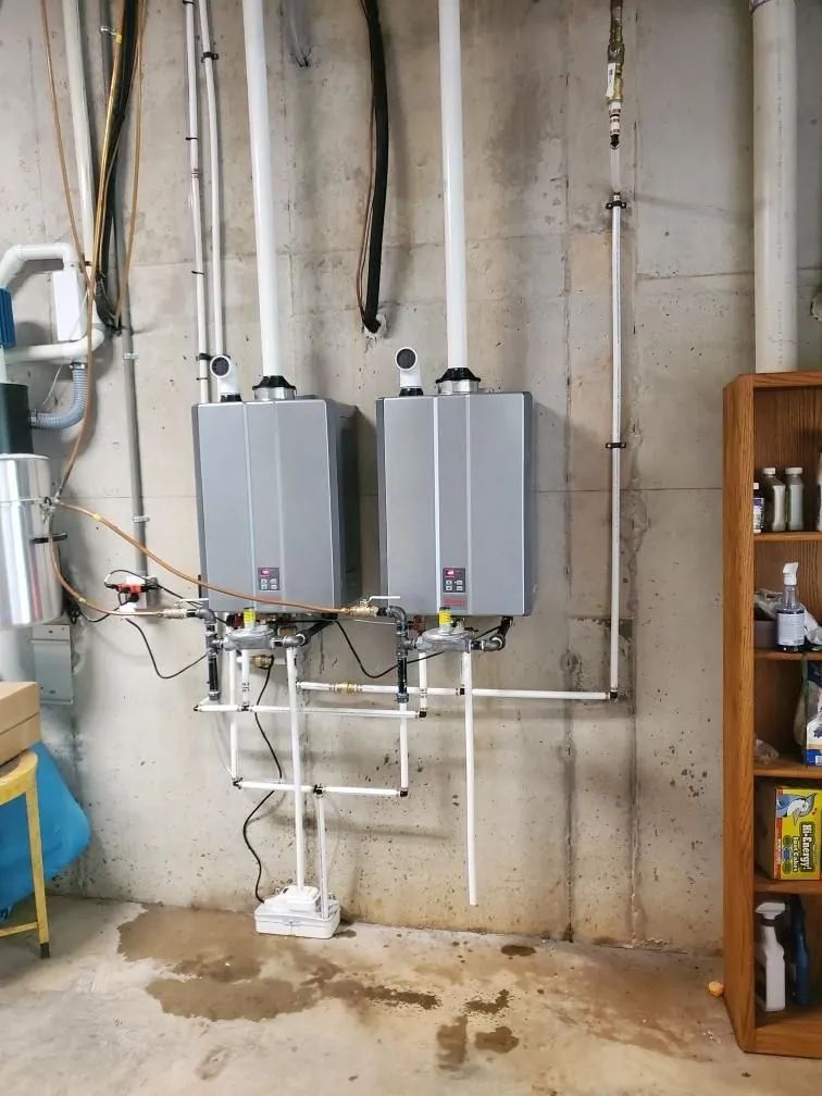 Two gray wall-mounted water heaters in a utility room with exposed pipes and a concrete floor.