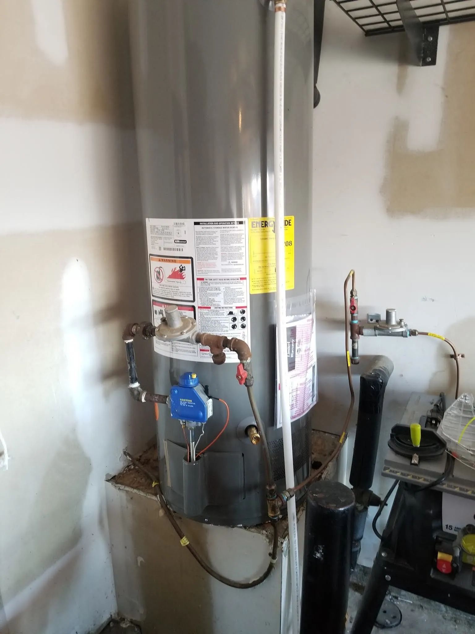 Water heater in a utility room with pipes, labels, and a blue control unit beside it.