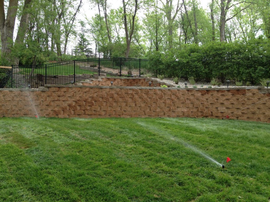 Lawn sprinkler spraying water on a green hillside below a brick retaining wall and wooded fence line.