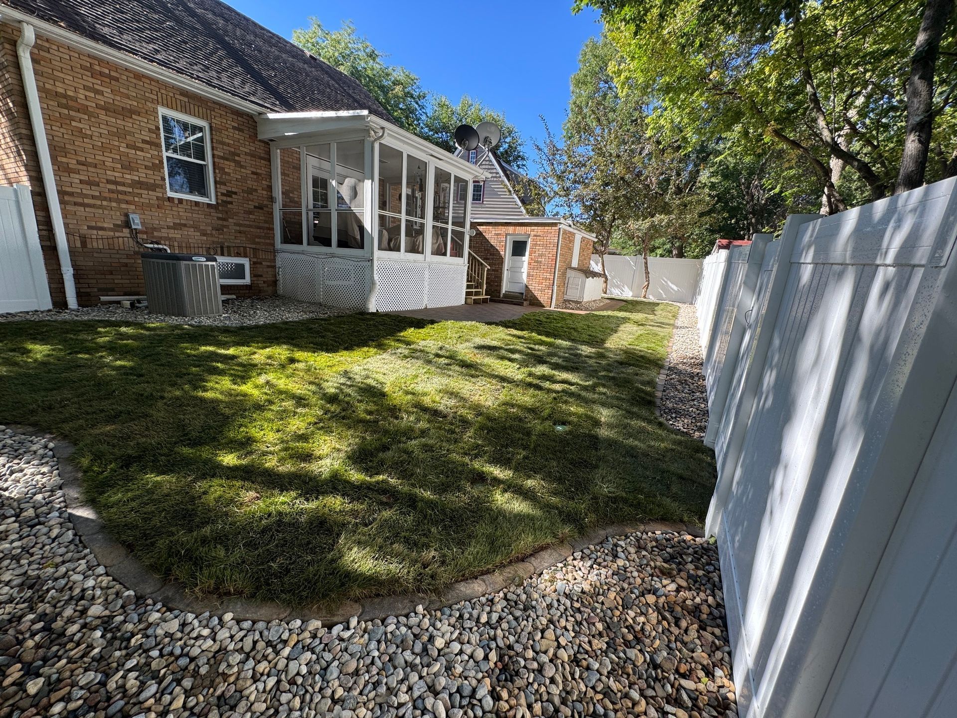 Backyard with sloped grass, gravel border, white fence, and brick house patio in sunlight