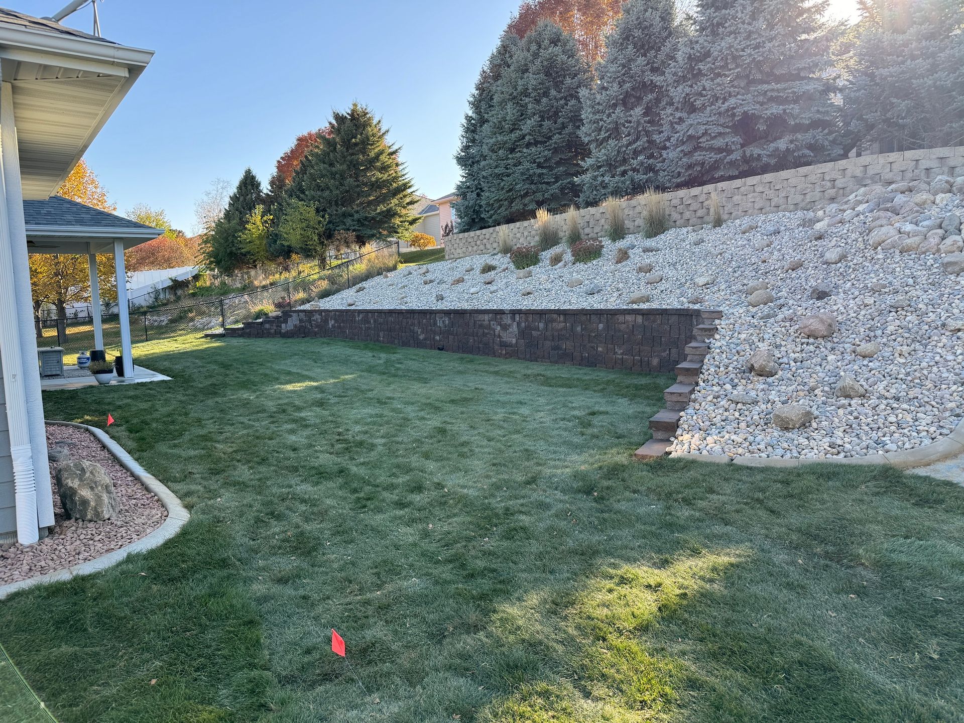 Sloped backyard lawn with a stone retaining wall, evergreen trees, and part of a house porch on the left.