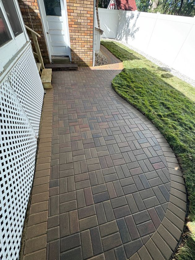 Brick-paved side yard path beside a house, bordered by white fence and green groundcover.